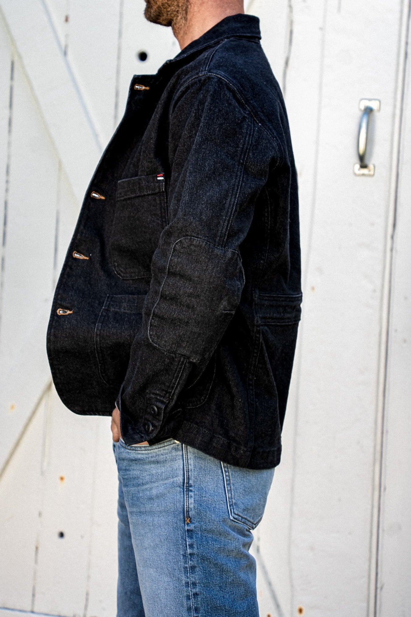 Side view of a men's black denim blazer showing elbow patch, buttoned cuff, and chest pocket, worn with light wash jeans.