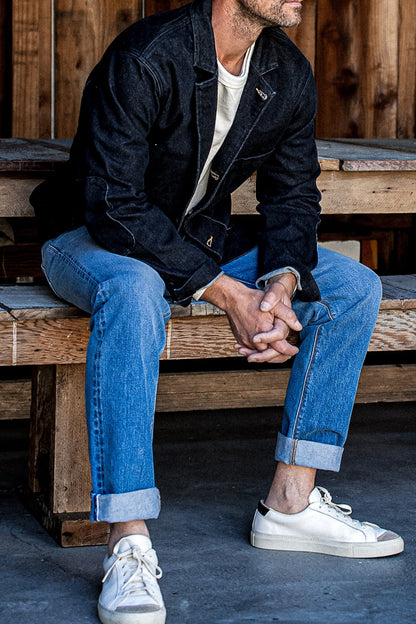 Man in a black denim chore blazer, cream tee, and cuffed blue jeans seated on a wooden bench.