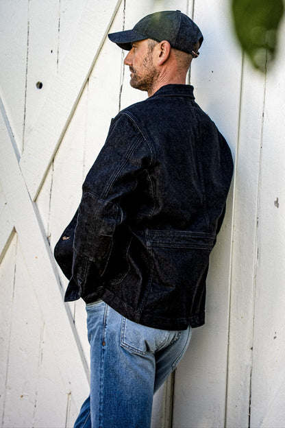 Rear side view of black denim chore blazer with elbow patch, worn against a white barn door.