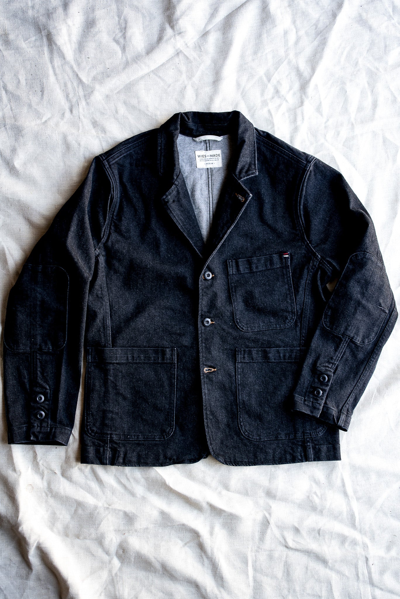 Black denim chore blazer laid flat on linen, showing patch pockets, elbow patches, and button front.