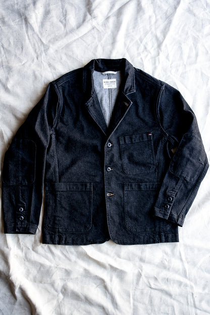 Black denim chore blazer laid flat on linen, showing patch pockets, elbow patches, and button front.