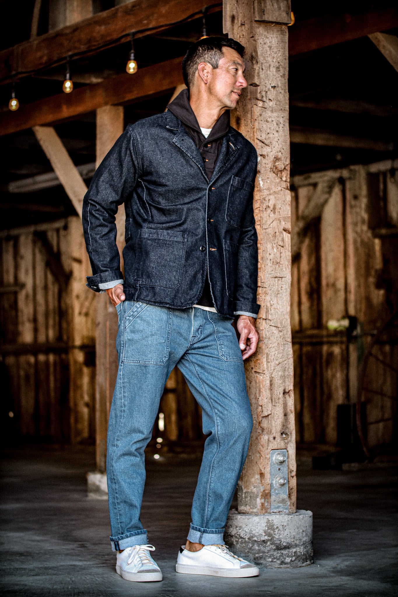 Man in a dark indigo denim chore blazer over a brown shirt and light-wash jeans, leaning on a wooden post.