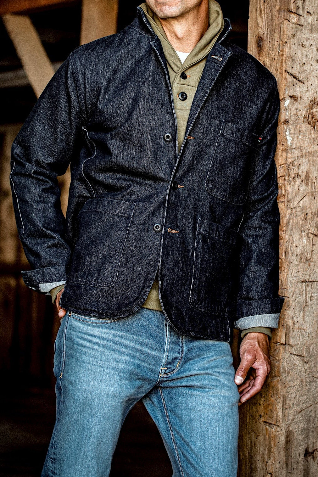 Man wearing dark indigo denim chore blazer with patch pockets over jeans, leaning on wood beam.