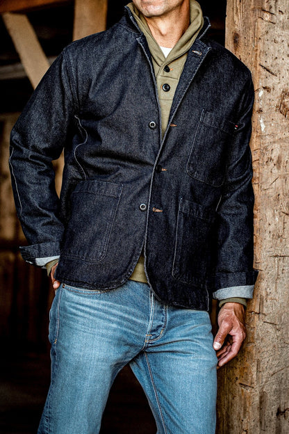 Man wearing dark indigo denim chore blazer with patch pockets over jeans, leaning on wood beam.