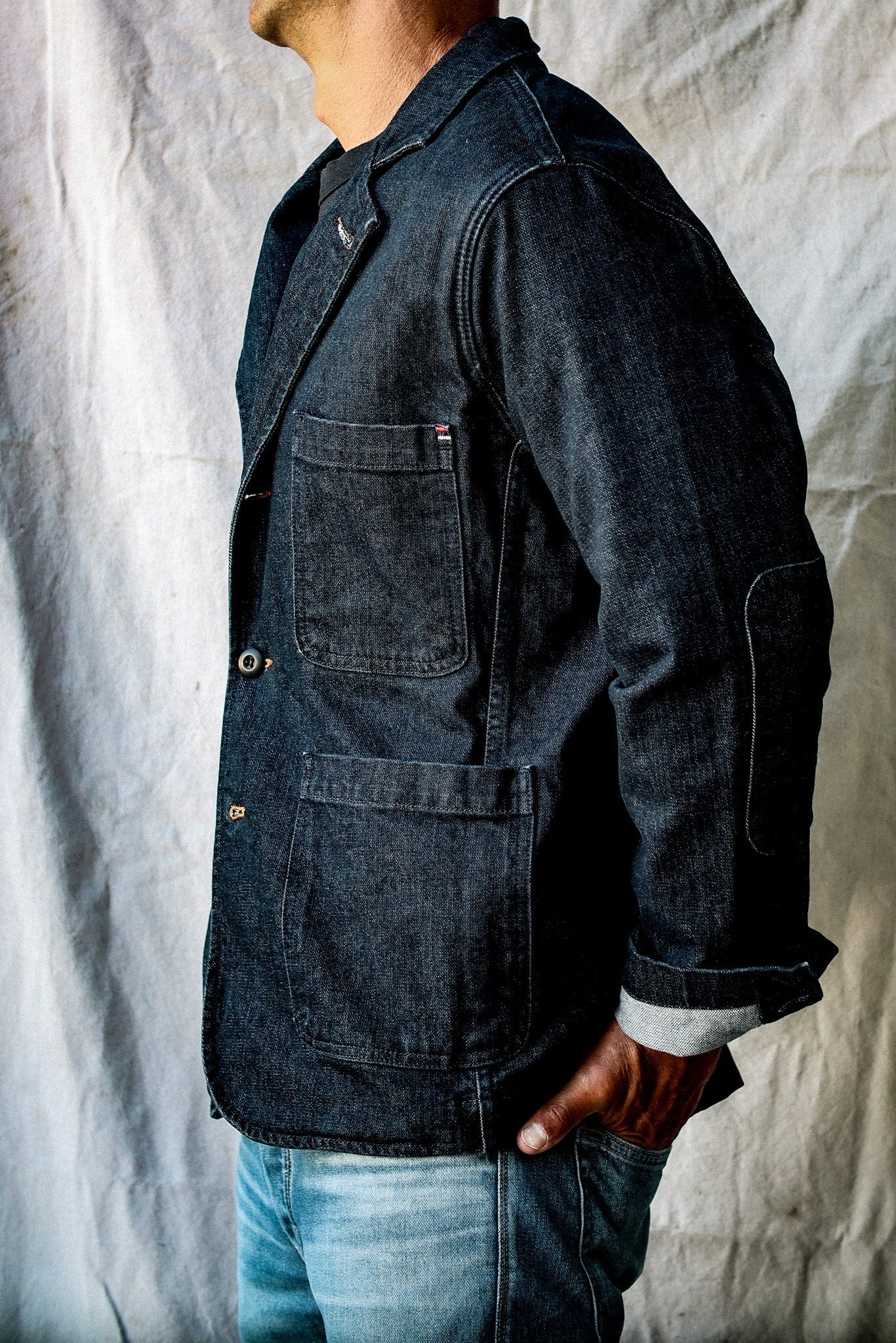 Side view of indigo denim chore blazer showing patch pockets, elbow patch and cuffed selvedge sleeve.