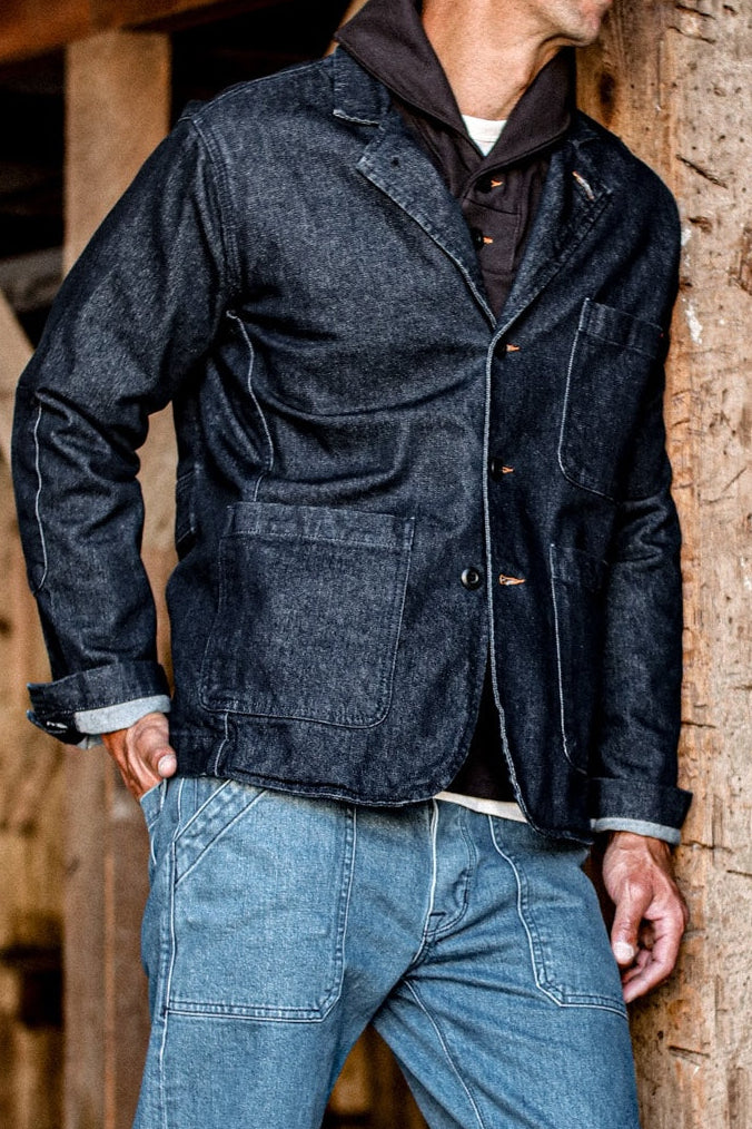 Person wearing indigo denim chore blazer with patch pockets, leaning against a wooden beam.