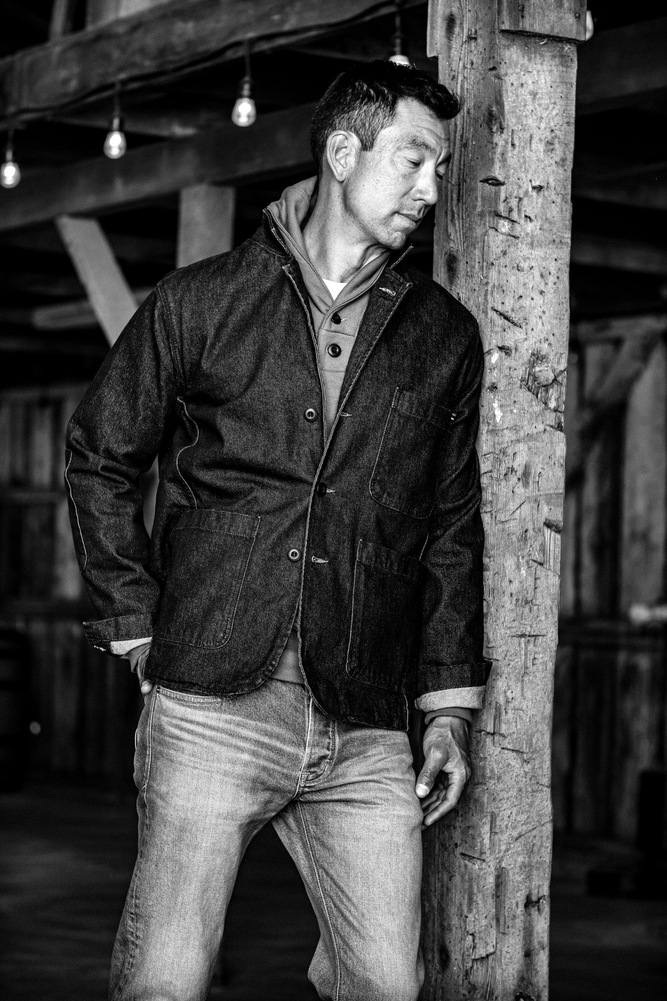Man in a denim chore blazer with patch pockets, leaning against a wooden post in a rustic barn.