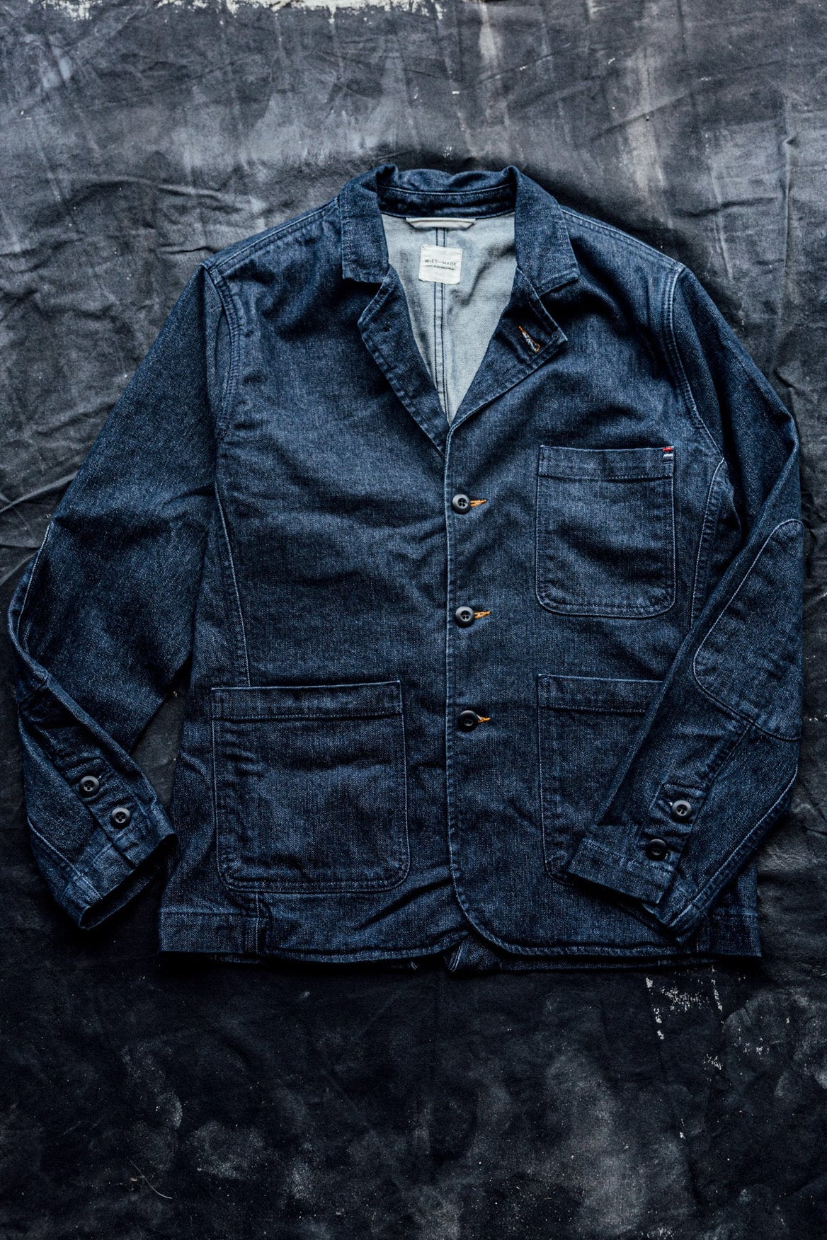 Men's denim blazer in indigo wash laid flat, showing notched lapels, patch pockets, and elbow patches.