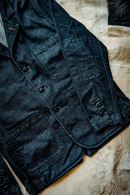 Ocean indigo denim chore blazer laid flat, showing patch pockets, black buttons, and tobacco stitching.
