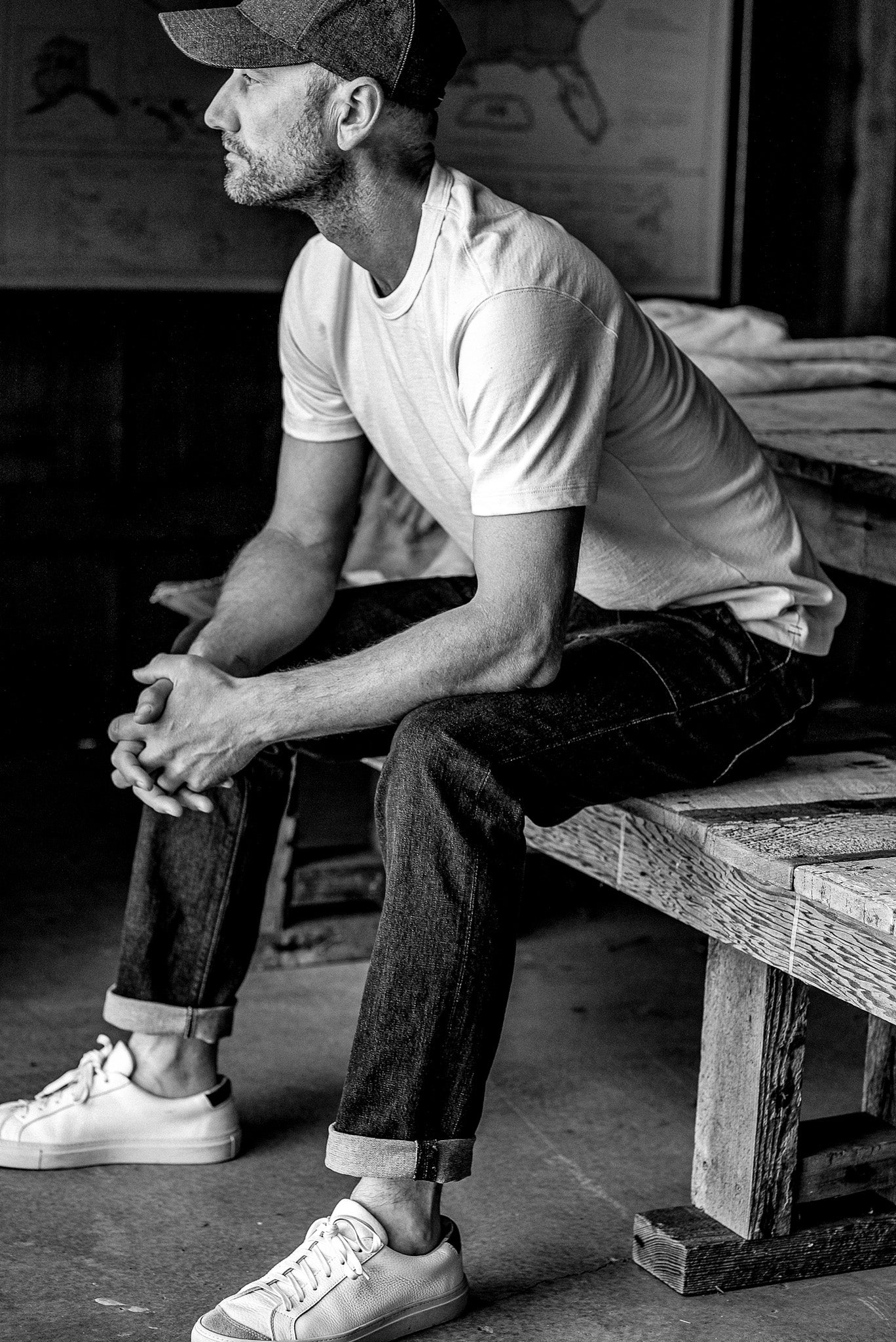 Man sitting on a wooden bench wearing cuffed selvedge denim jeans, a white tee, and sneakers.