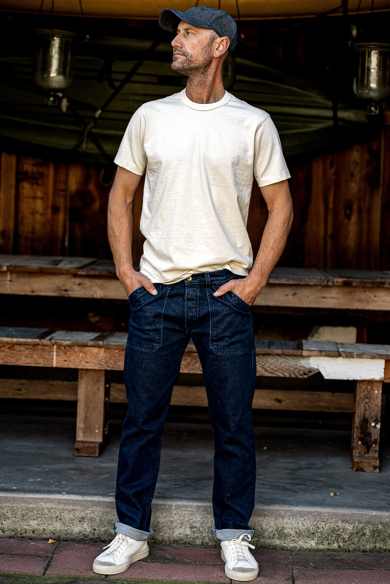 Man wearing Scout military-style denim jeans in ocean wash with cream tee and white sneakers outside a wooden cabin.