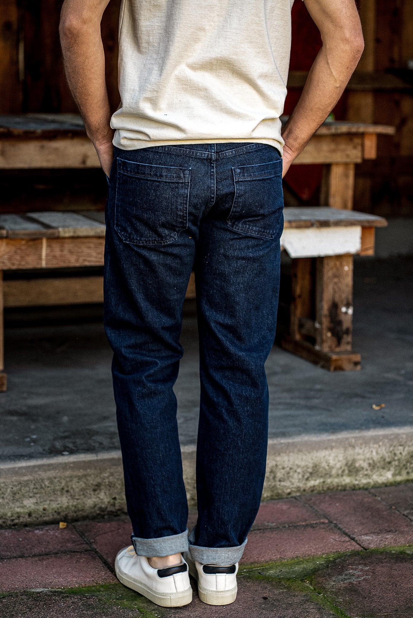 Back view of men's dark ocean wash denim jeans with cuffed hems, paired with a cream tee and white sneakers.