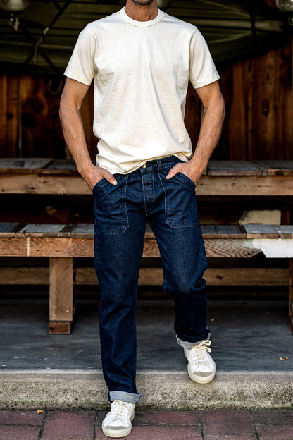 Man wearing dark ocean wash denim military pants with front utility pockets, cuffed hems, cream tee, and white sneakers.