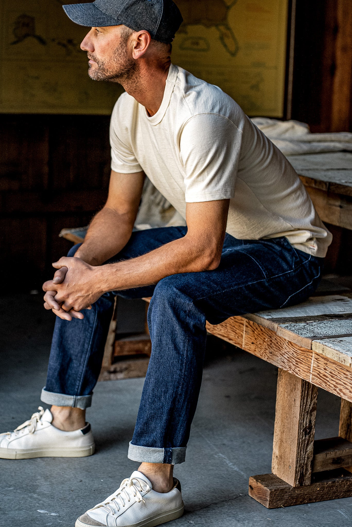 Man in cream tee and cuffed Scout ocean wash selvedge denim jeans seated on a weathered wood bench.