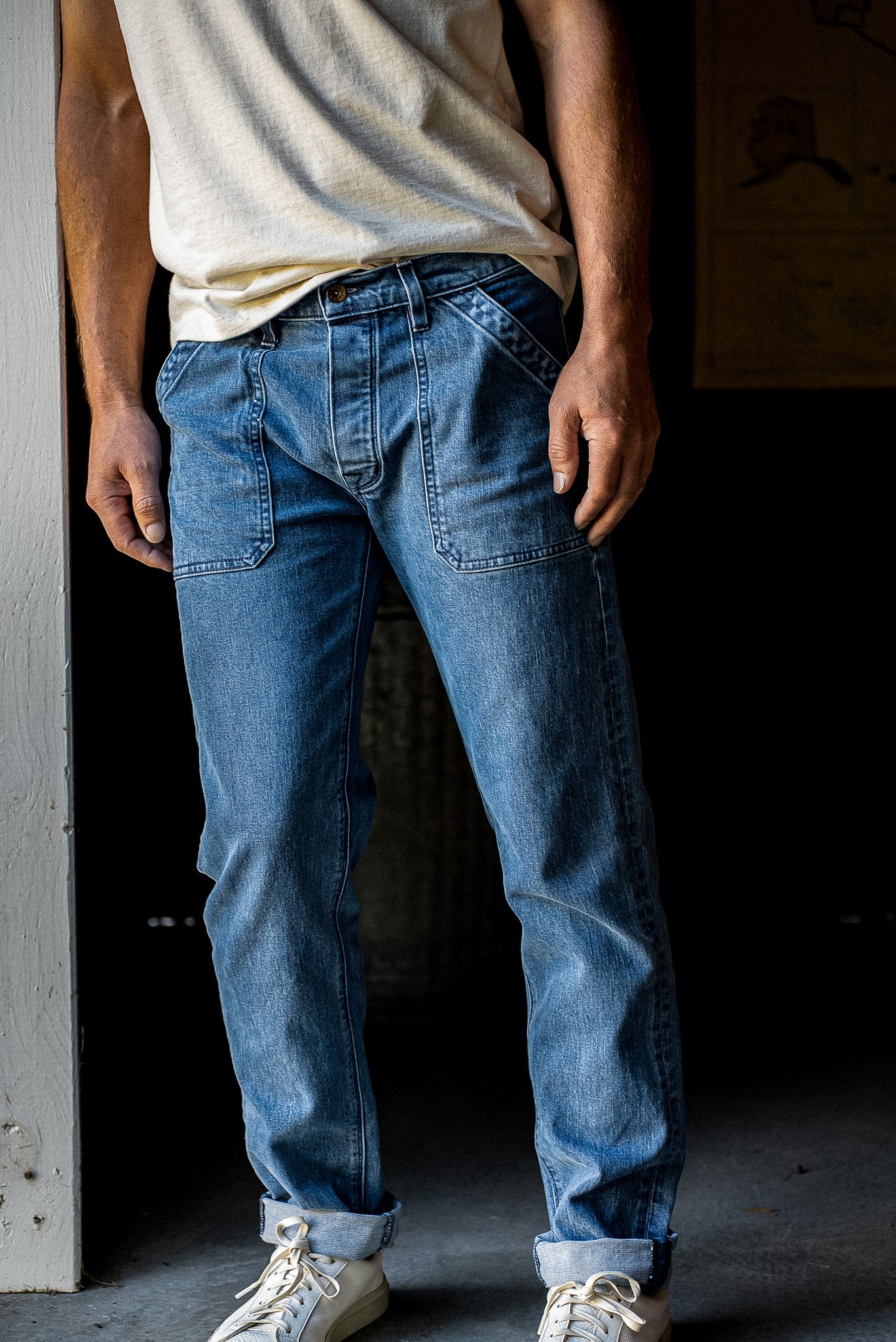 Man wearing salt-wash Scout military denim jeans with patch pockets, cuffed hems, and white sneakers.