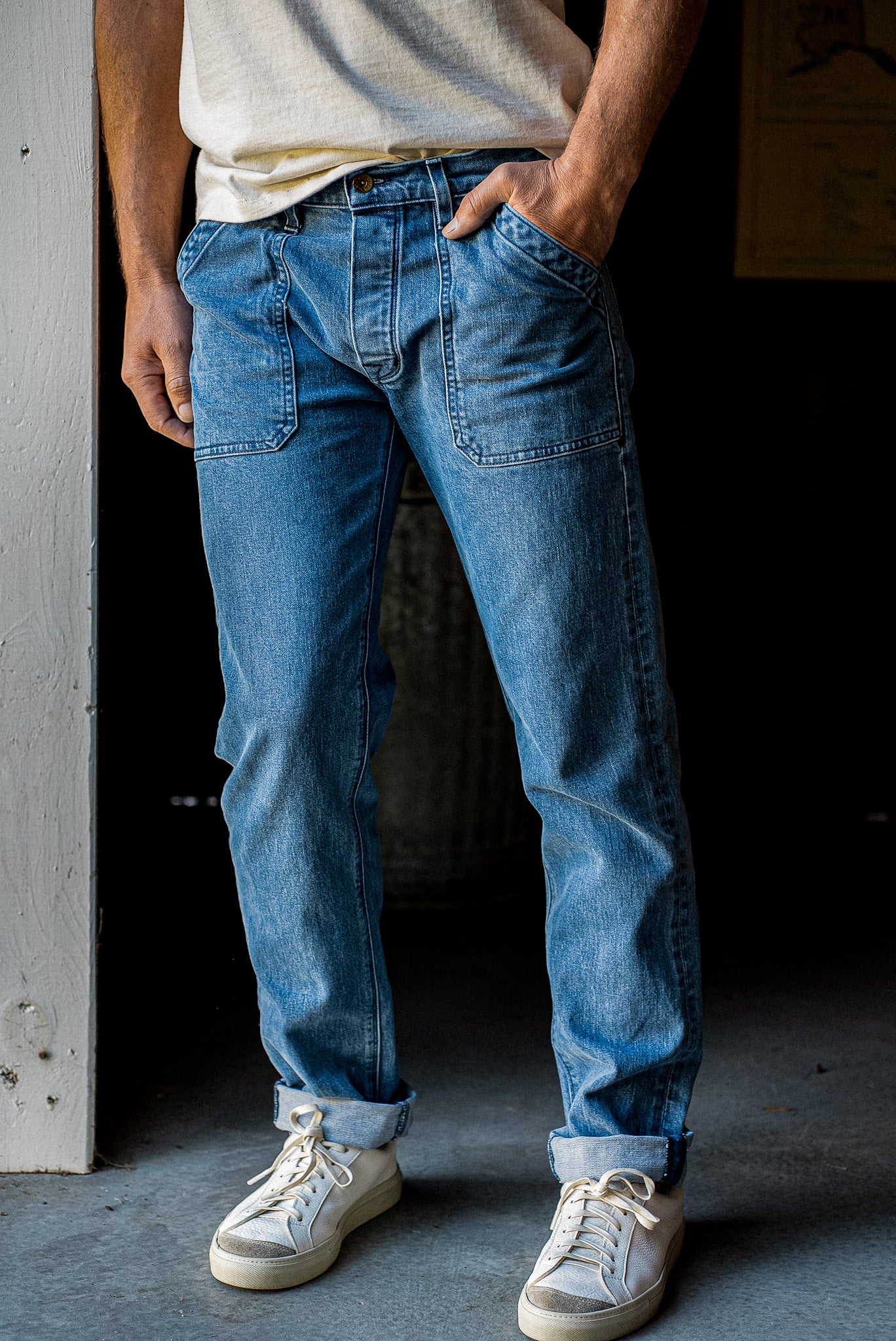 Salt-wash men's denim jeans with military patch pockets, cuffed hems, paired with a cream tee and white sneakers.