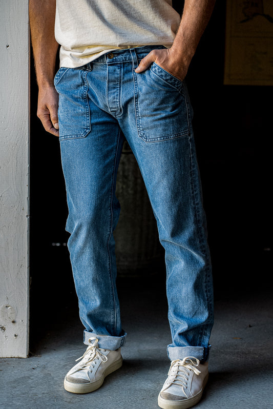 Salt-wash men's denim jeans with military patch pockets, cuffed hems, paired with a cream tee and white sneakers.