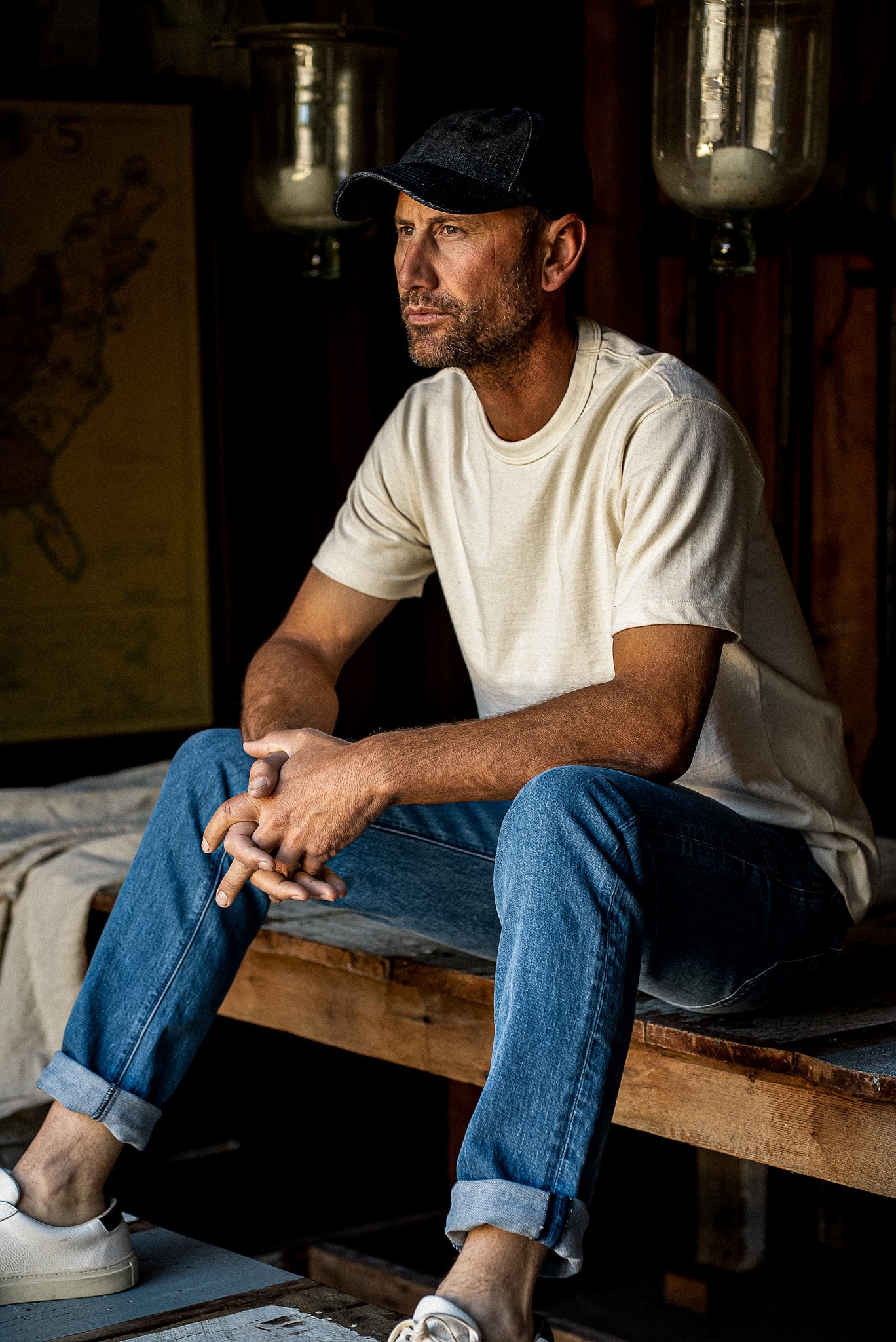 Man in cream tee and black cap sitting on a wooden bench wearing salt-wash Scout straight-leg denim jeans with cuffed hems.