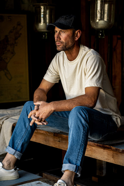 Man in cream tee and black cap sitting on a wooden bench wearing salt-wash Scout straight-leg denim jeans with cuffed hems.