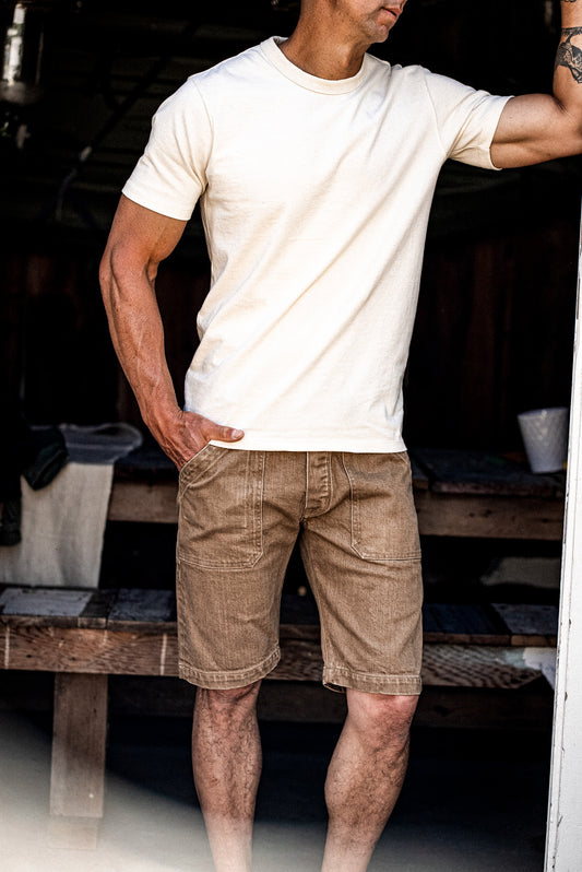 Man in wheat-colored denim military shorts and cream tee leaning against a weathered workshop doorway.