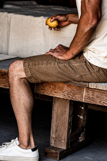 Man in wheat-colored denim military shorts seated on a wooden bench with white sneakers.