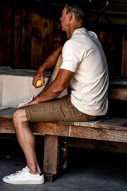 Man in wheat denim military shorts and cream polo seated on a rustic wooden bench.