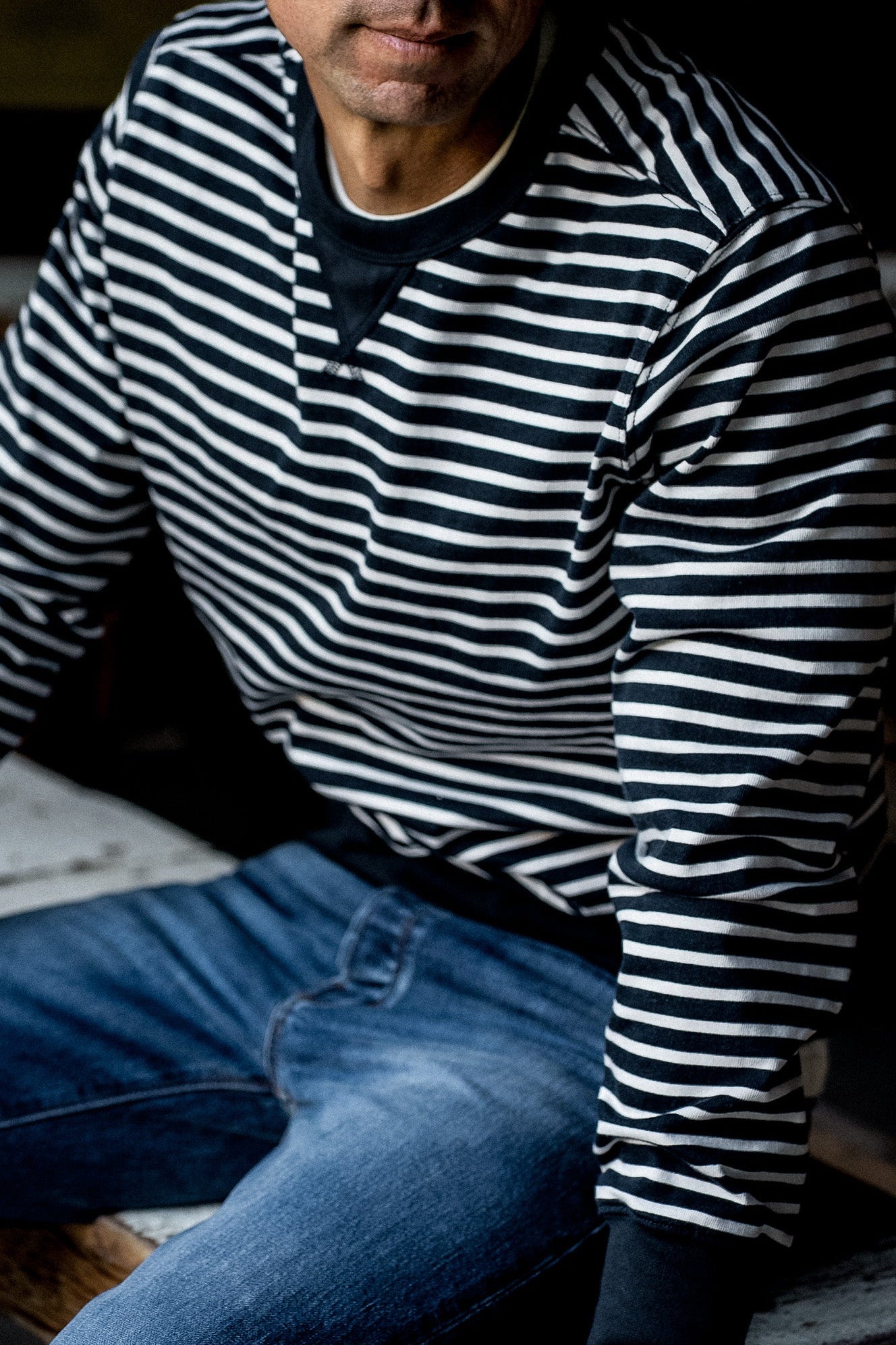 Man wearing a navy and white striped rugby jersey pullover with jeans, seated.
