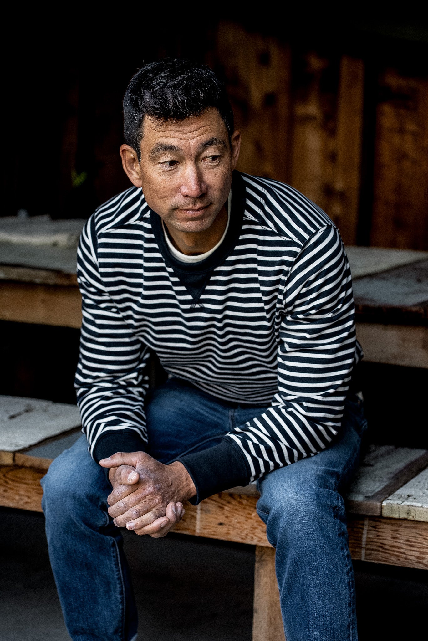Man in a black and white striped rugby jersey pullover and jeans seated on a wooden bench.