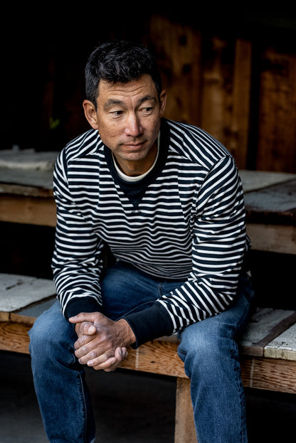 Man in a black and white striped rugby jersey pullover and jeans seated on a wooden bench.