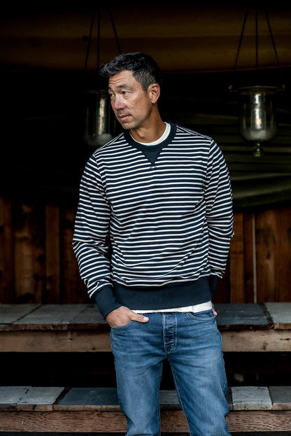 Man in a navy and cream striped Stinson rugby jersey pullover and blue jeans on a wooden porch.
