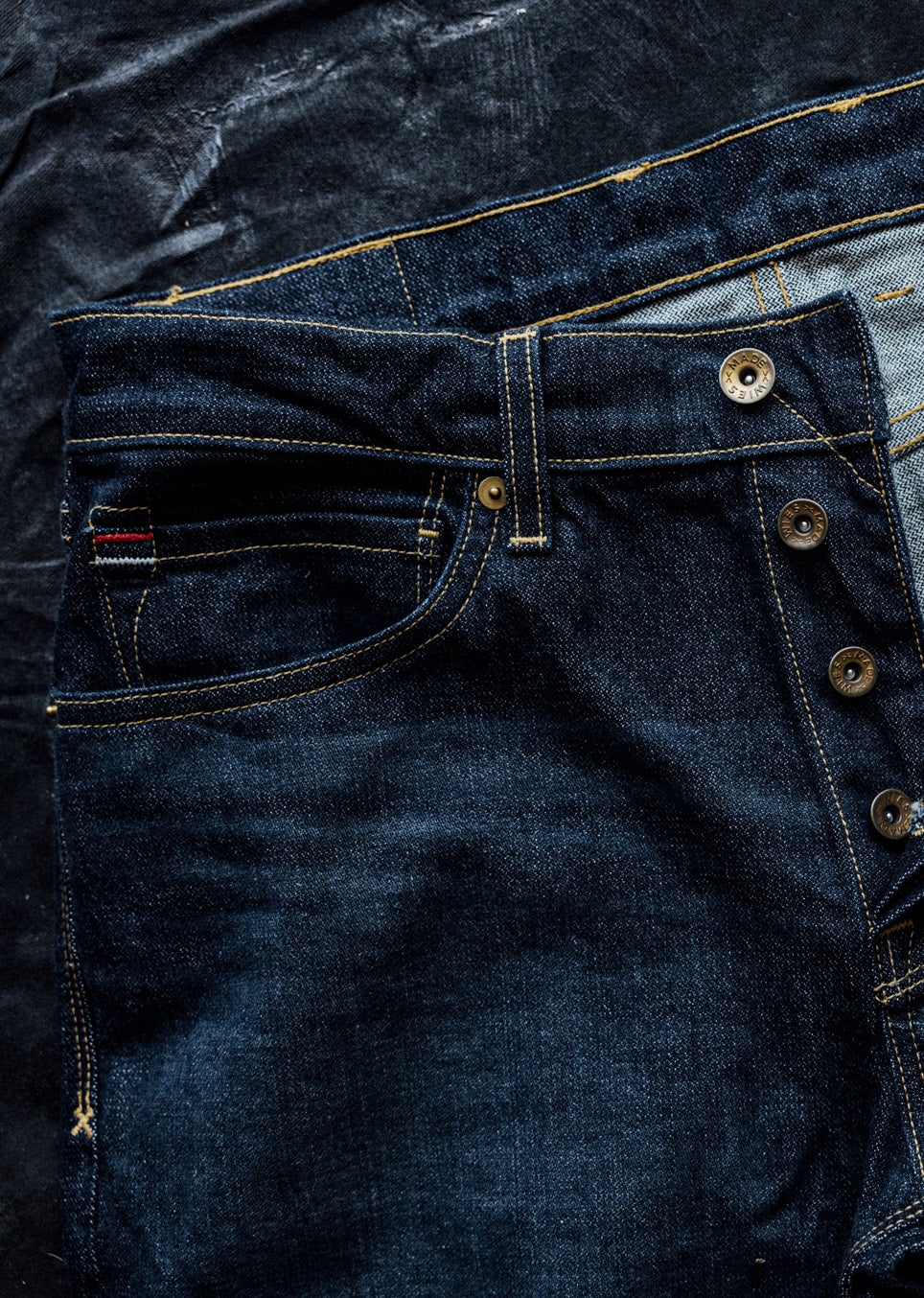 Close-up of dark ocean wash denim jeans showing brass button fly, contrast stitching, and selvedge detail.