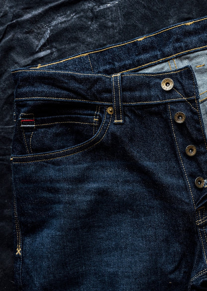 Close-up of dark ocean wash denim jeans showing brass button fly, contrast stitching, and selvedge detail.