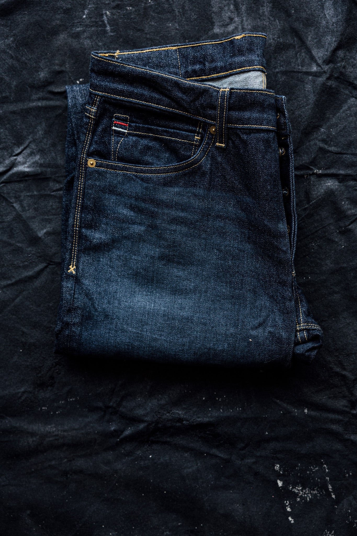 Men's slim straight jeans in dark denim, folded neatly on a black textured background.