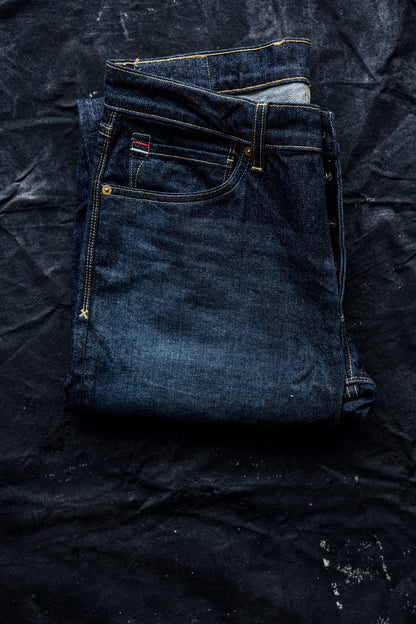 Folded dark ocean wash slim fit jeans showing contrast gold stitching, brass rivets, and red selvedge tab on a black canvas.