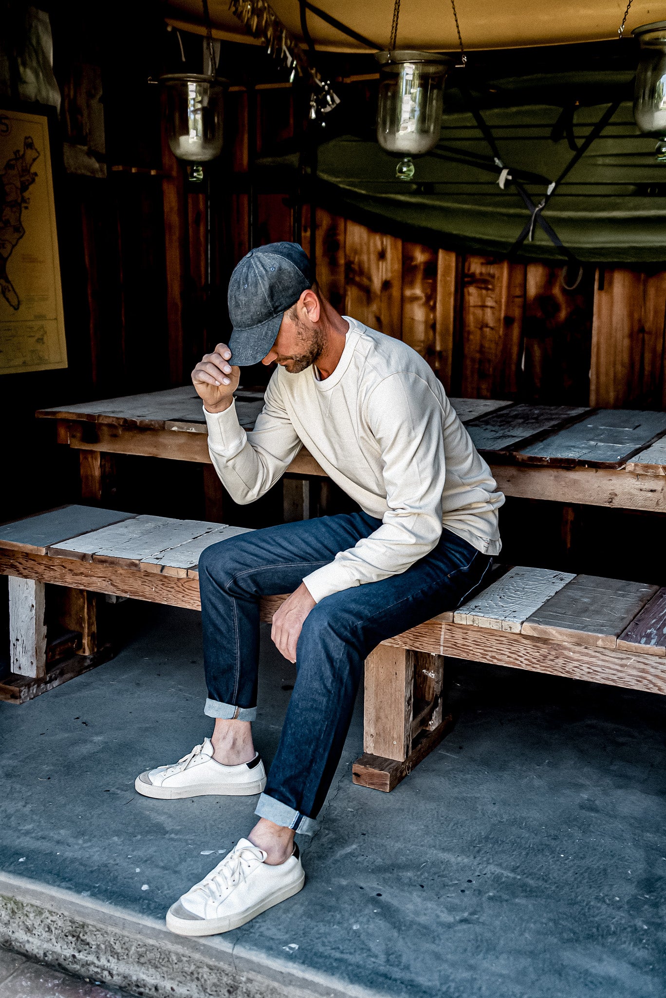Man in ocean wash straight slim fit jeans with cuffed hem, seated on a wooden bench in a rustic shed.