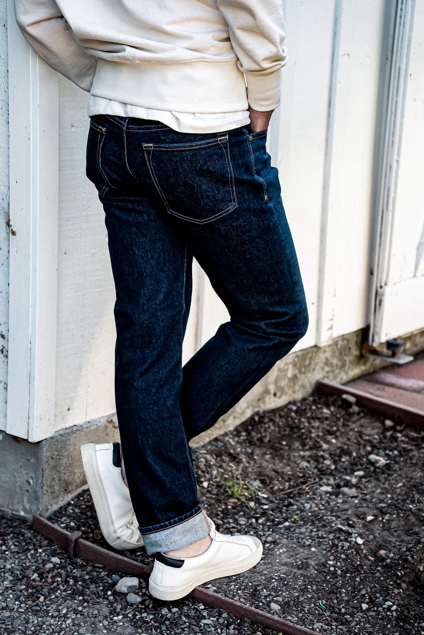 Back view of dark ocean wash straight slim fit jeans with cuffed hems, worn with cream sweatshirt and white sneakers.