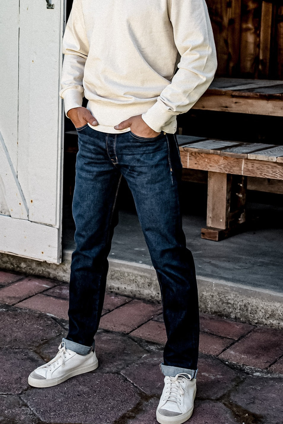 Model in ocean wash straight slim jeans with cuffed hems, cream sweatshirt, and white sneakers on a brick sidewalk.