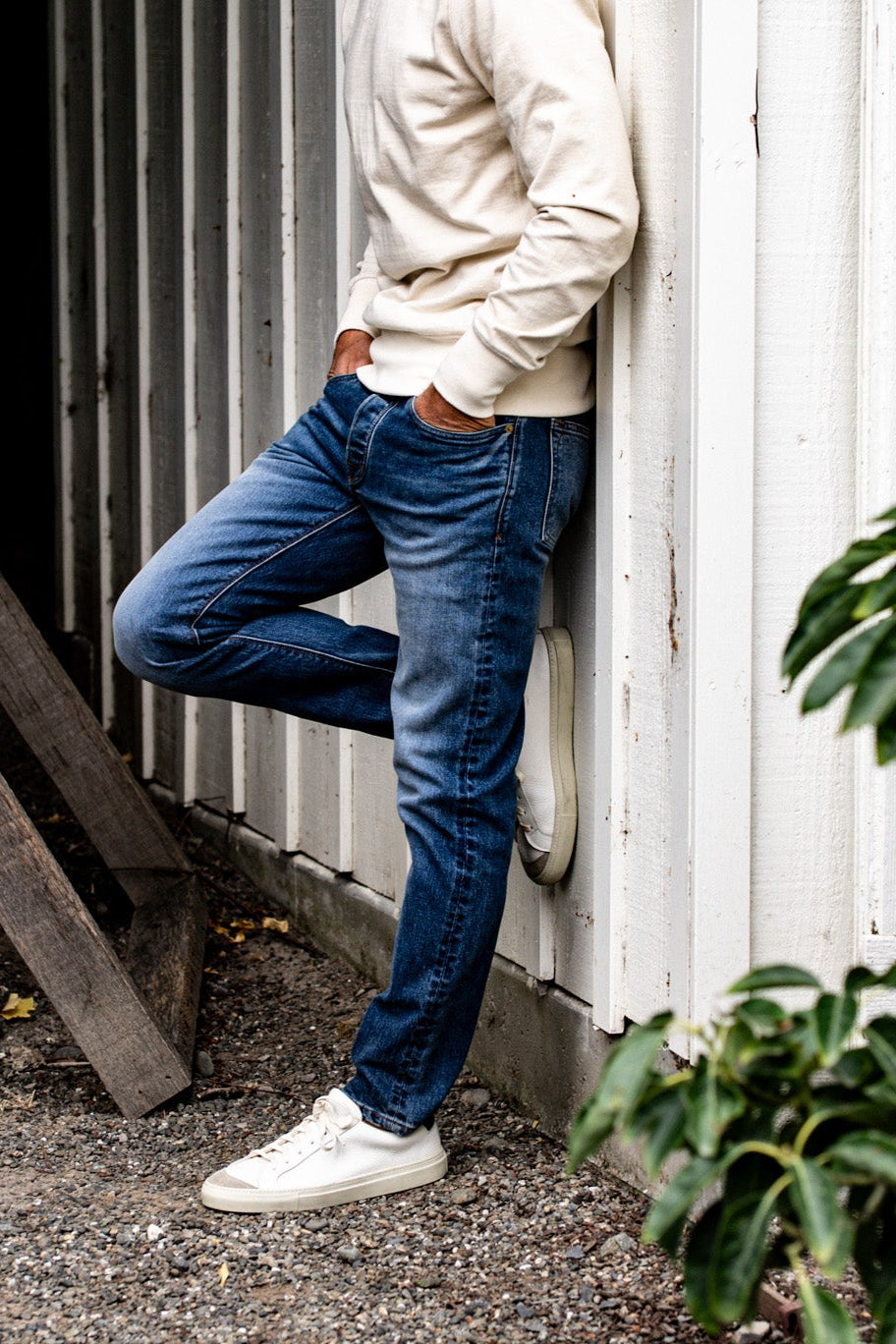 Man leaning against a white wall wearing men's premium denim jeans and a beige hoodie