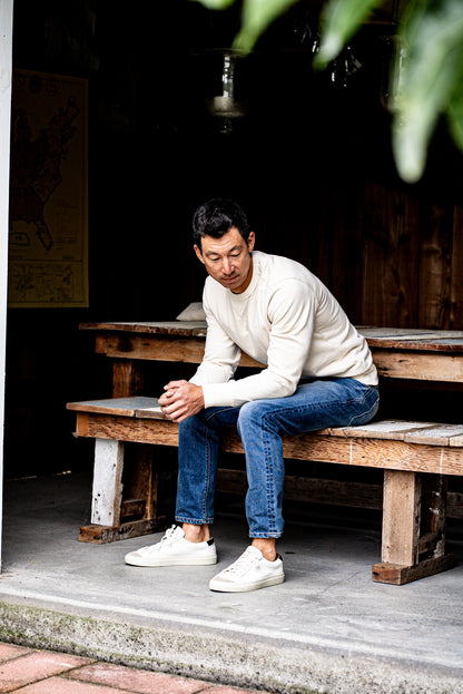 Man sitting on a wooden bench outdoors wearing slim fit jeans made in USA and a white sweater