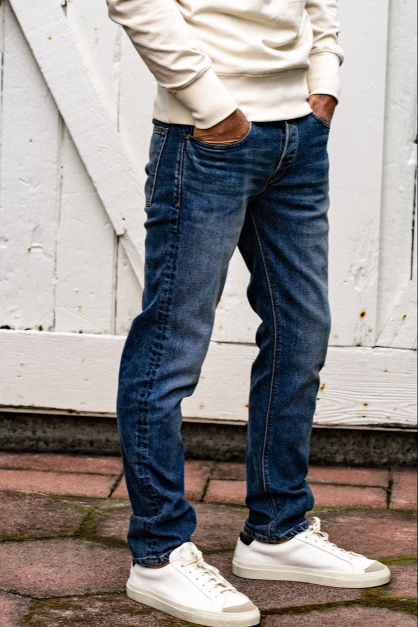 Man wearing men's straight slim fit jeans made in USA and white sneakers, standing against a wooden wall