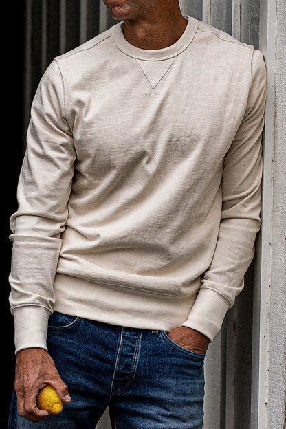 Natural cream crewneck rugby jersey pullover worn with dark denim jeans against a corrugated metal wall.