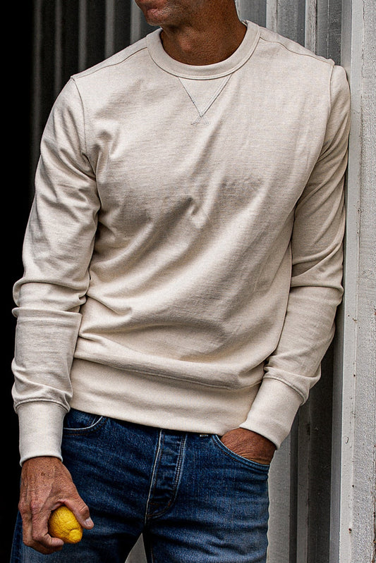 Natural cream crewneck rugby jersey pullover worn with dark denim jeans against a corrugated metal wall.