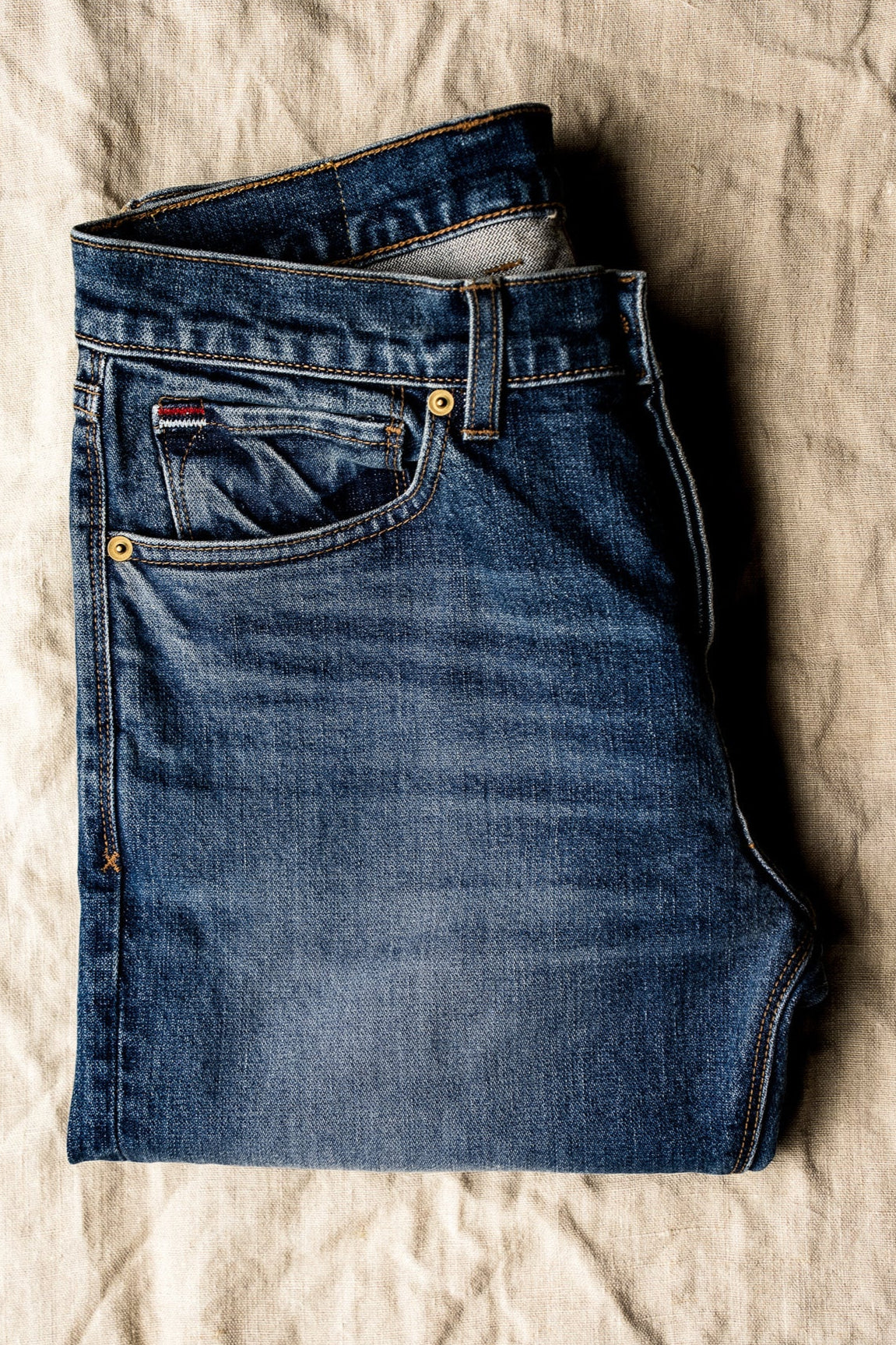 Men’s designer blue jeans folded neatly on a neutral fabric backdrop, featuring a midweight denim texture, classic five-pocket design, and soft fading for a clean, versatile aesthetic