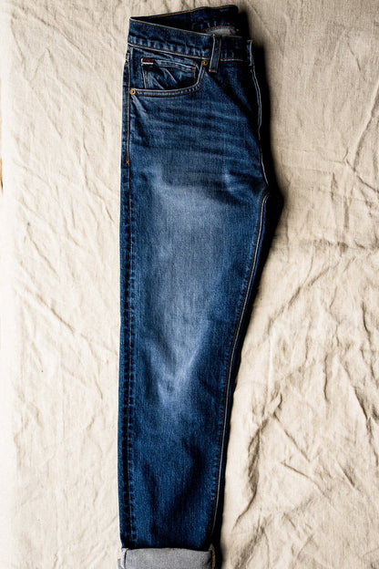 Men’s designer blue jeans neatly folded on a textured fabric surface, showcasing a tapered leg, medium wash finish, and classic five-pocket detailing with refined stitching