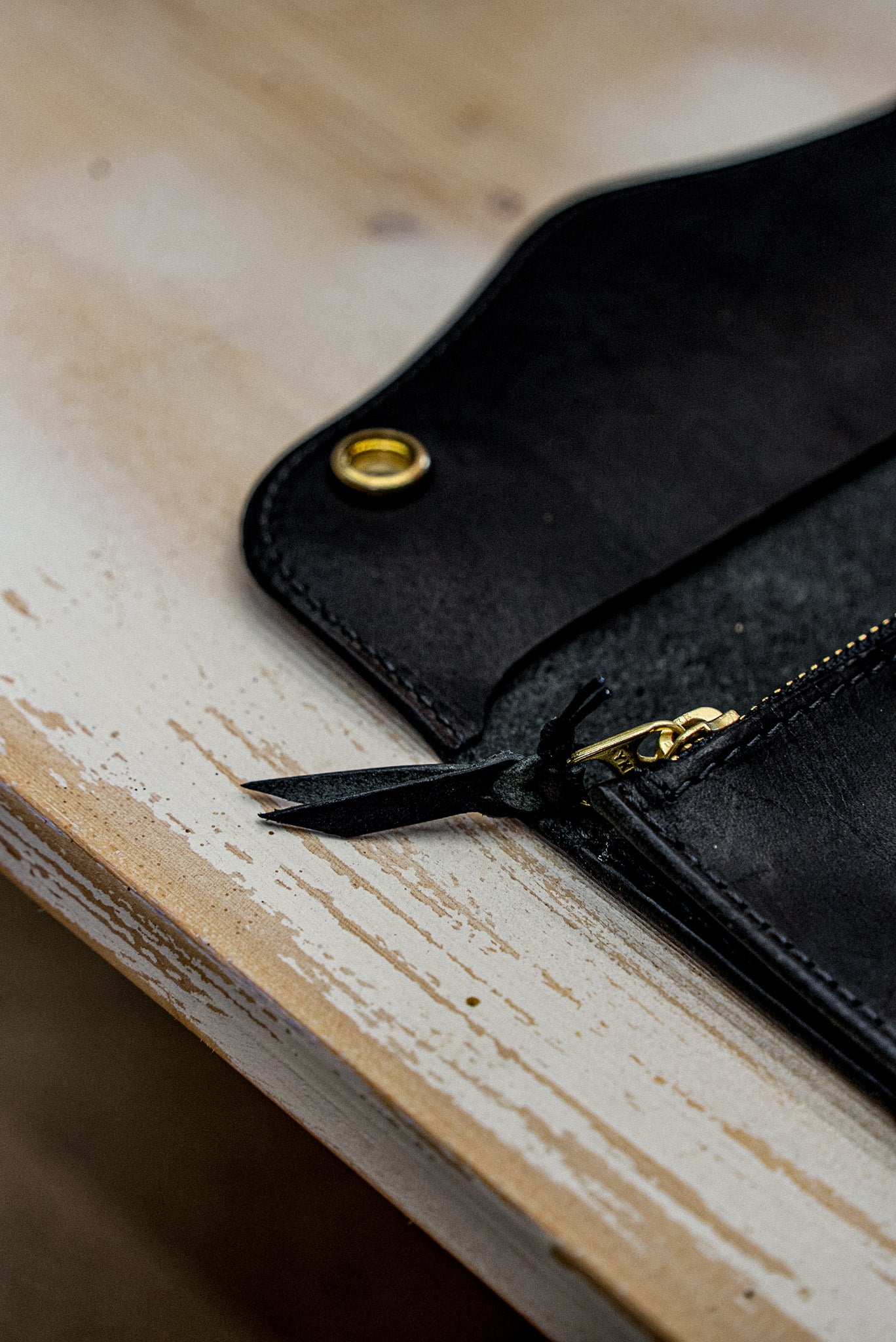 Black Horween leather Roper wallet with brass zipper and rivet on a rustic workbench.