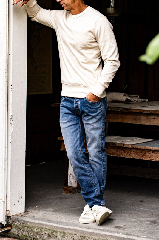 Men’s premium denim jeans styled with an off-white sweatshirt and sneakers, featuring a medium blue wash, straight leg cut, and subtle fading for a polished everyday look