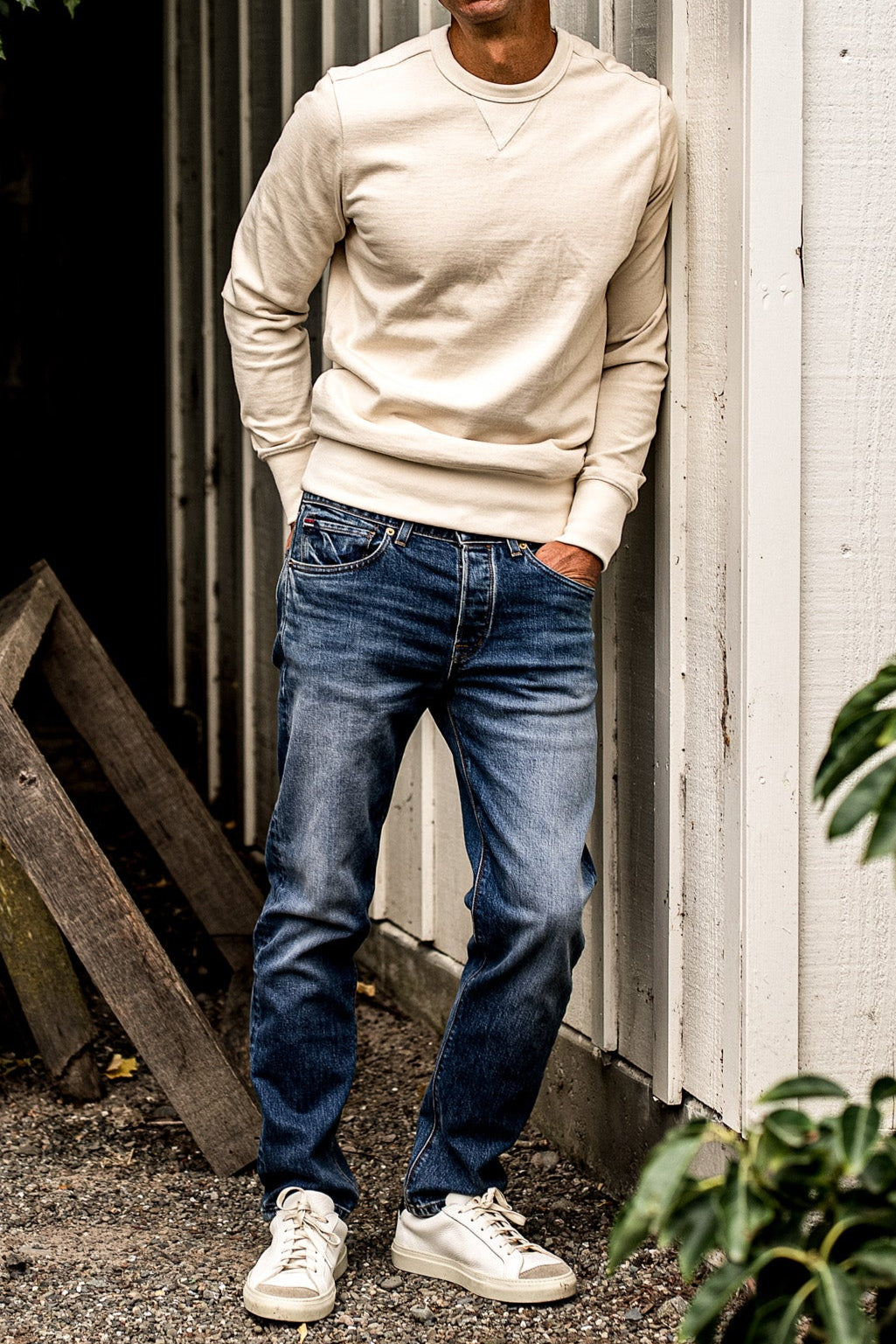 Denim designer jeans for men paired with a cream sweatshirt and sneakers, featuring a slim straight fit, medium blue wash, and light fading for a clean, modern aesthetic