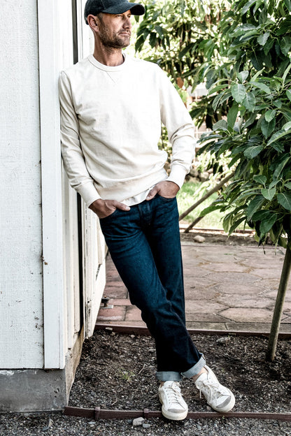 Man in dark ocean wash American made denim jeans with cuffed hems, cream sweatshirt, and white sneakers leaning against a.