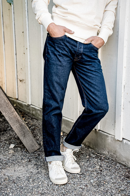 Model in dark ocean wash American made denim jeans with cuffed hems, paired with cream sweatshirt and white sneakers against.