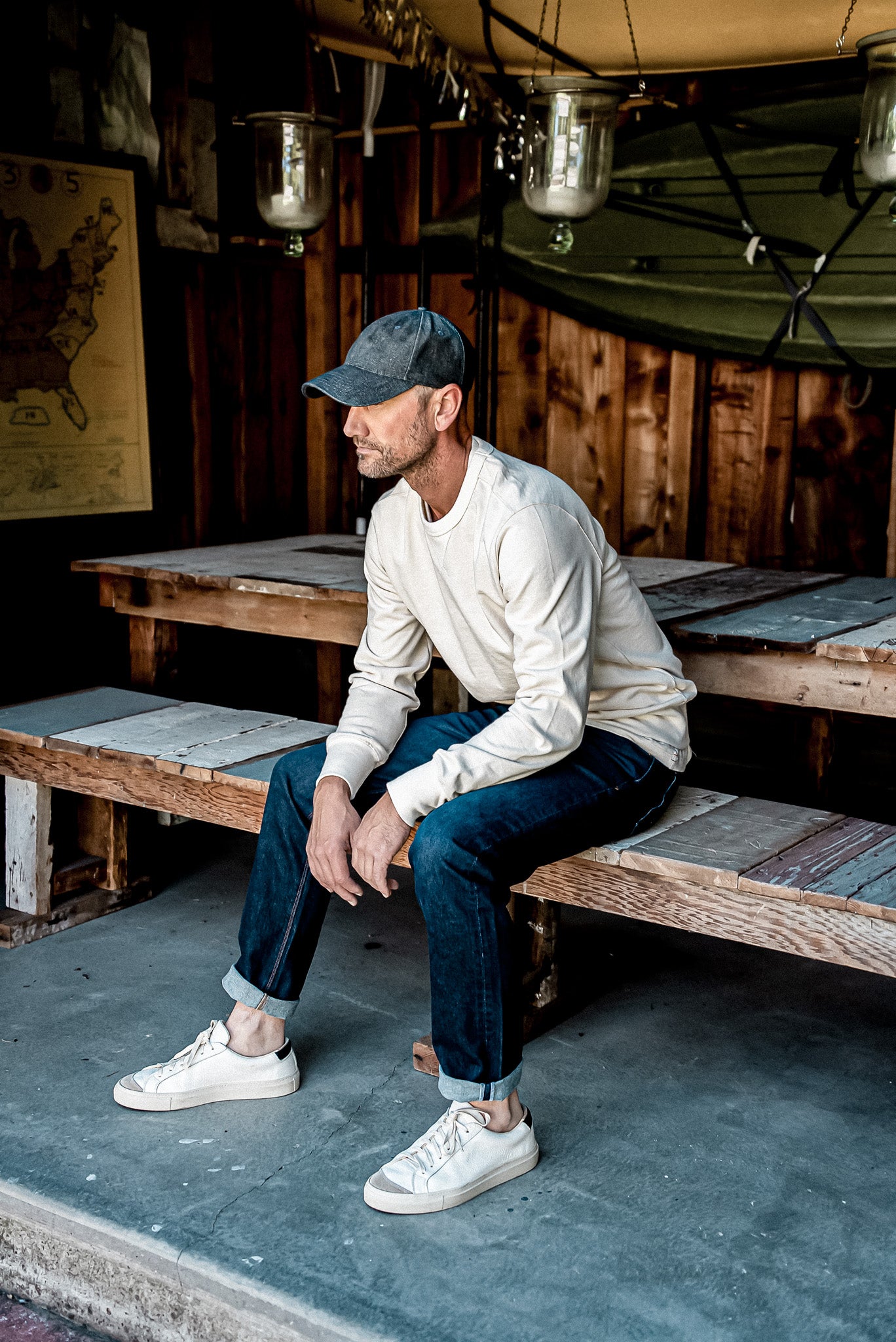 Man in cuffed dark ocean wash American made denim jeans, cream sweatshirt and cap, seated on a wooden bench.